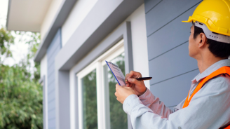 An inspector wearing a hard hat and vest takes notes as he examines the outside of a house.